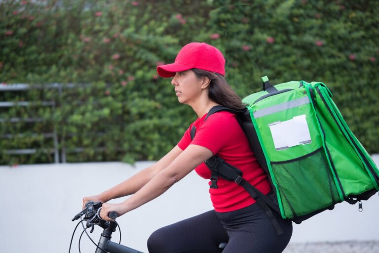 A Delivery Woman in Red Cap and Shirt Carrying an Insulated Bag while Riding a Bicycle