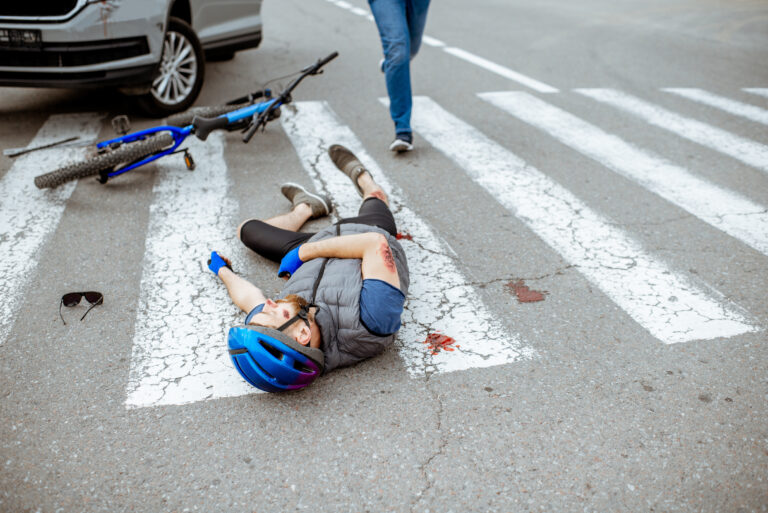 Road accident with injured cyclist lying on the pedestrian crossing near the broken bicycle and car driver running on the background